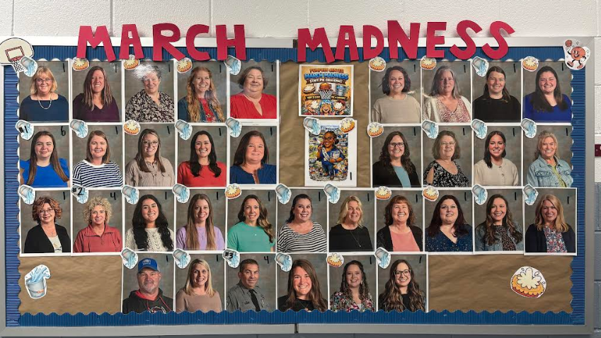 A "March Madness" themed bulletin board featuring a grid of staff portrait photographs. The top of the board has large red cutout letters spelling "MARCH MADNESS." The board is decorated with small graphics of basketballs being dunked and buckets of water being poured. In the center, a flyer titled "PUMPKIN CENTER MARCH MADNESS STAFF PIE CHALLENGE!" explains a competition where the top 16 winners get a pie in the face. Below that is a cartoon illustration of a staff member. The grid consists of four rows of staff photos, each labeled with a small number in the corner. Some photos have a "pie" graphic or a "water bucket" graphic attached to them, indicating their status in the challenge.