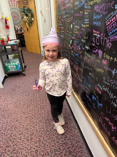 A young girl with long blonde hair, wearing a white long-sleeve shirt with purple patterns and black leggings, stands in a school hallway. She is wearing a pink and white paper crown that says, "I passed my 2nd grade sight words!" and features a "Hooray" graphic with a smiley face. She holds a purple marker and stands next to a large black chalkboard wall covered in colorful handwritten names. The background shows a rolling cart and an open office door with a floral wreath.