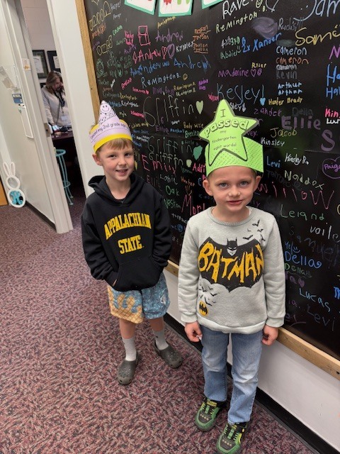 Two young boys stand side-by-side in front of a large black chalkboard wall covered in colorful handwritten student names. The boy on the left wears a black Appalachian State hoodie and a yellow paper crown that says, "I passed my 2nd grade sight words!" The boy on the right wears a grey Batman sweatshirt and a green star-shaped paper crown that reads, "I passed my Kindergarten sight words!" They are both smiling for the photo in a school hallway.