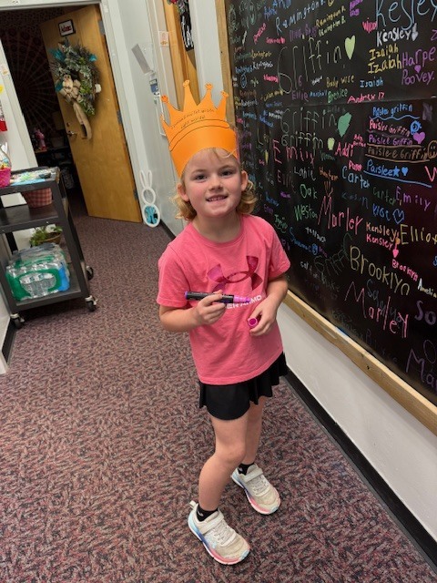 A young girl in a pink t-shirt and black skirt stands in a school hallway, smiling while wearing an orange paper crown that says, "I passed my 1st grade sight words!" She is holding a pink marker and standing next to a large black chalkboard wall filled with student names written in various colors. In the background, a rolling cart and an open doorway are visible.
