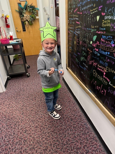 A young boy smiling brightly while wearing a green paper star crown that reads, "I passed my Kindergarten sight words!" He is standing in a school hallway next to a large black chalkboard wall covered in colorful handwritten names. He holds a marker in his hands, and in the background, a rolling cart with a pink basket and a decorated door are visible.