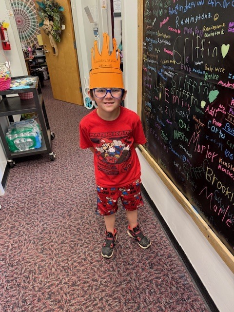 A young boy with blue glasses wearing a red Mario Kart t-shirt and matching Mario shorts stands in a school hallway. He is wearing an orange paper crown that says, "I passed my 1st grade sight words!" He is standing next to a black chalkboard wall covered in colorful handwritten names. In the background, a rolling cart with cases of water and a door with a wreath are visible.