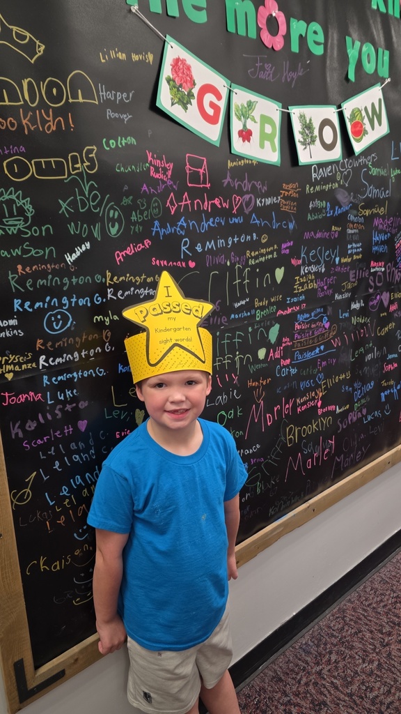 A young boy in a blue t-shirt and tan shorts stands in front of a large black chalkboard wall. He is wearing a yellow paper crown shaped like a star that reads, "I passed my Kindergarten sight words!" The chalkboard behind him is covered in colorful handwritten names of students and drawings. Above the boy, a banner spells out "GROW" with illustrations of plants, and the wall features the phrase "the more you" at the top.