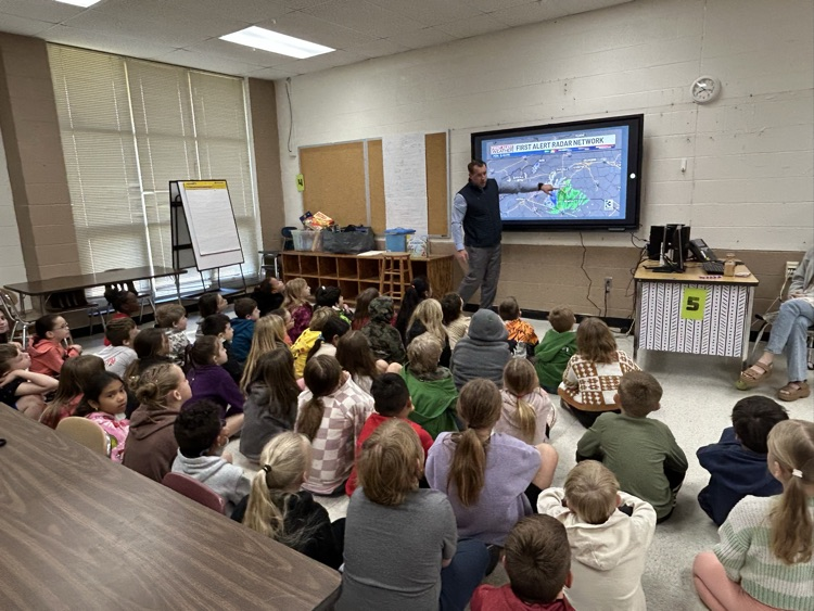 A male presenter stands in a classroom pointing to a large digital screen displaying a "First Alert Radar Network" weather map with green and blue radar echoes over a regional map. A large group of elementary-aged students sits on the floor facing the screen, listening to the presentation. The classroom features cinderblock walls, a wooden cubby storage unit, a white clock on the wall, and a table to the right with a sign labeled with the number 5.