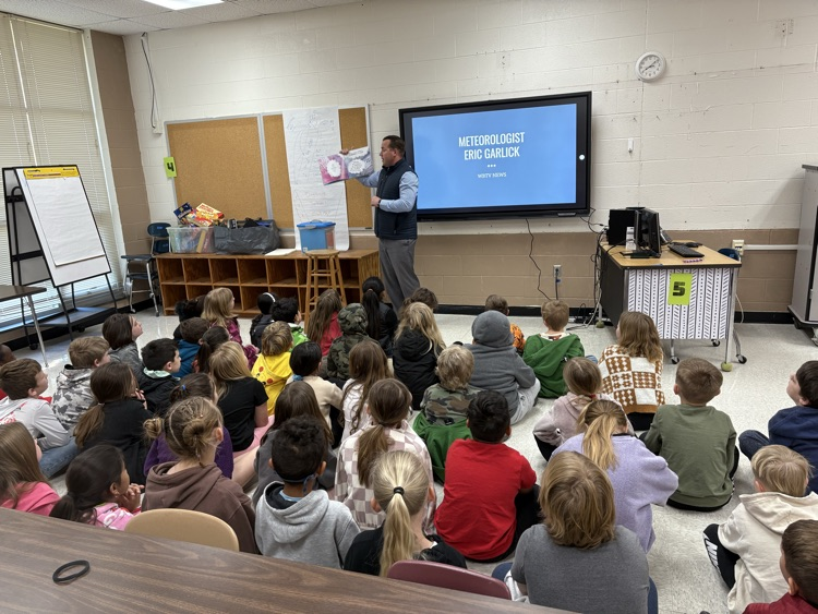 Meteorologist Eric Garlick stands in a classroom holding up a children's book to a large group of elementary students seated on the floor. Behind him, a digital screen displays the text Meteorologist Eric WBTV News. The students are looking toward the presenter in a classroom setting with cinderblock walls and educational materials. 