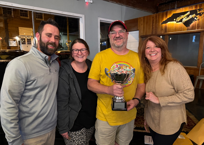 Four adults pose together, smiling, with one person in a yellow shirt holding a trophy. They stand inside the same venue, suggesting a team or award recognition moment.