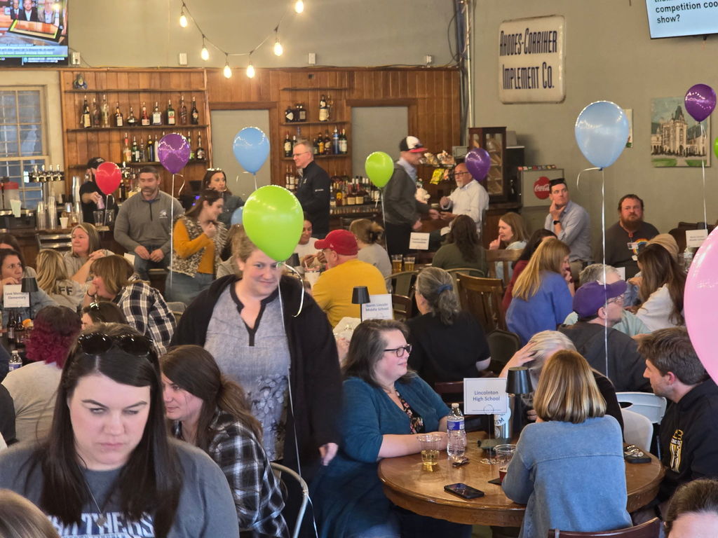 A large group of adults gathered inside a warm, wood-paneled venue, seated at tables and standing near a bar area. Colorful balloons float above tables, and people are talking, eating, and socializing in a lively atmosphere.