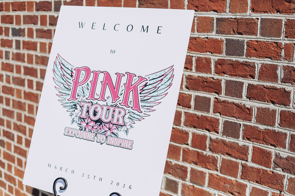 A sign on an easel reads “Welcome to Pink Tour: Exposure to Inspire” with the date March 25th, 2026, displayed in front of a brick wall.