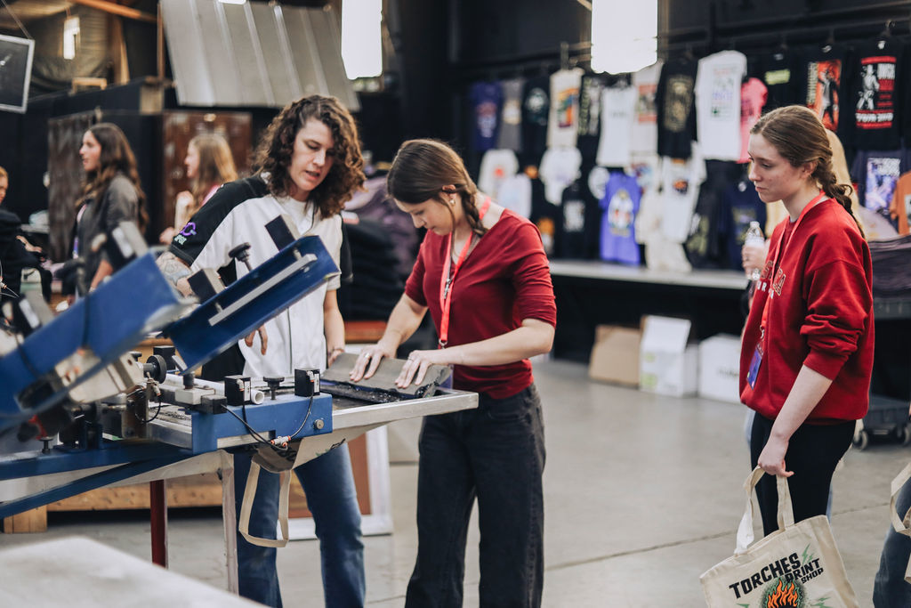 A group of high school students observe and participate in a hands-on demonstration with a printing press inside a manufacturing facility, while an adult guides them through the process.
