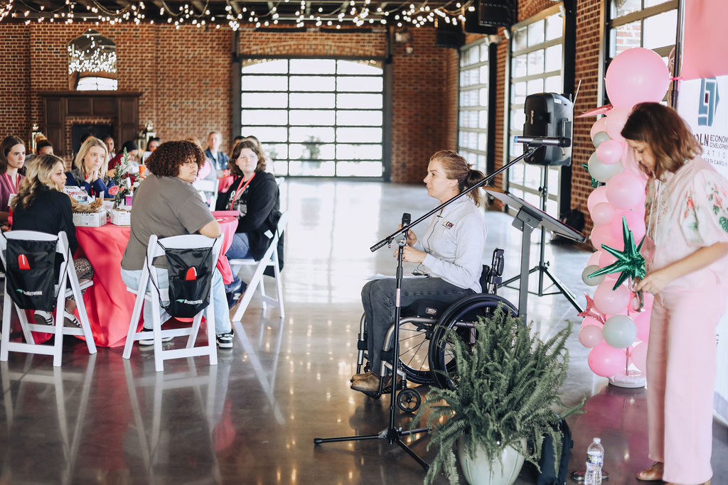 A woman in a wheelchair speaks into a microphone at a luncheon event, while high school students seated at tables listen attentively in a large, decorated venue with string lights and pink accents.