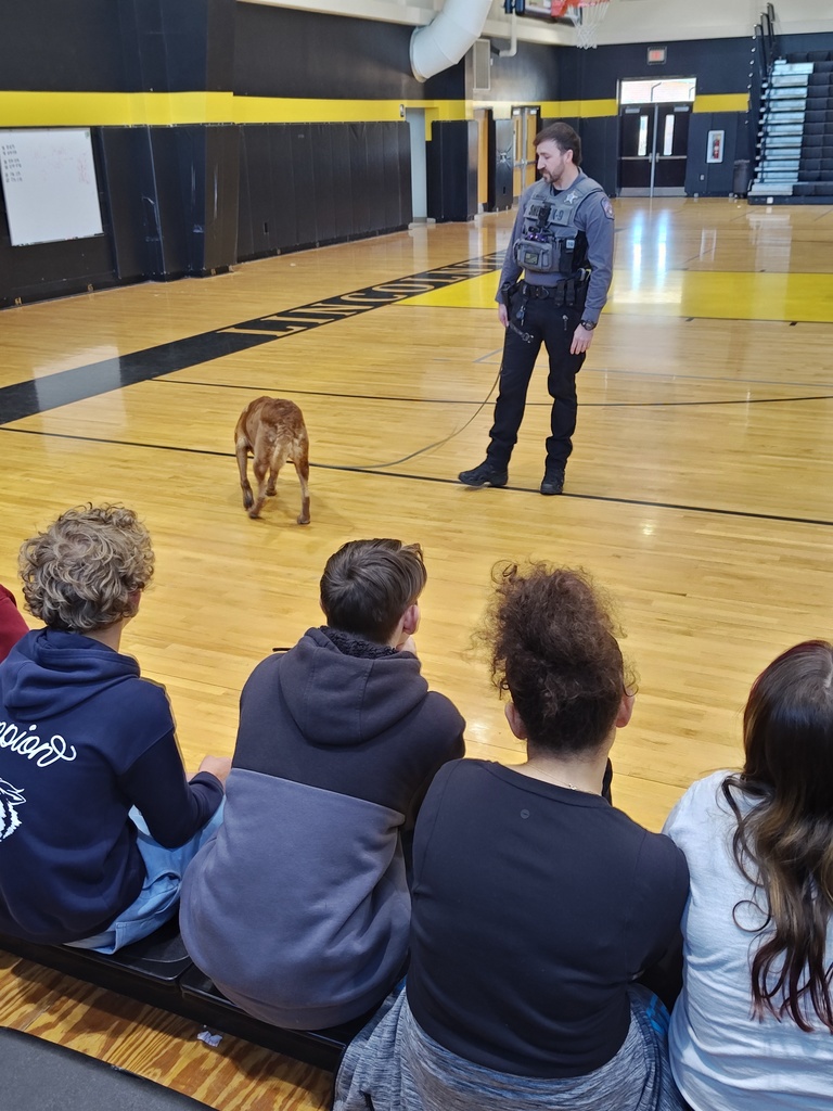 Officer Wilson stands in a school gymnasium with his K-9, Marty, a brown dog. Students sit in the foreground watching as the officer and K-9 prepare for a demonstration on the wooden basketball court.
