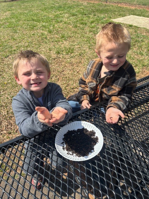 Students exploring soil and worms Two young boys sit at a black metal mesh picnic table outdoors on a sunny day. In the center of the table is a white paper plate filled with dark soil.  Key Details Interaction: The boy on the left, wearing a grey zip-up jacket, holds his hands out toward the camera showing small worms. The boy on the right, wearing a brown and black plaid flannel shirt, looks down at a worm in his own hand with a smile.  Setting: They are positioned on a grassy area with a concrete walkway visible in the background.  Activity: The image captures a hands-on science or nature lesson, likely related to gardening or biology.