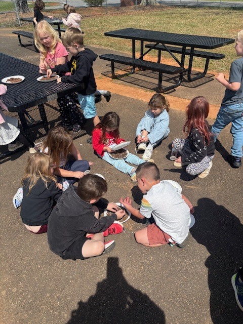 A classroom of students exploring soil outdoors A high-angle shot showing a large group of elementary students sitting in a circle on a paved outdoor area next to a grassy field. They are fully immersed in a hands-on science lesson on a bright, sunny day.  Key Details Activity: Nearly a dozen students are kneeling or sitting on the ground, each focused on white paper plates filled with soil, mulch, and organic matter. They are using their hands and small tools to sort through and examine the samples.  Environment: The scene is set outdoors at school, with green grass and backpacks visible in the background. The students are dressed in colorful casual clothes (t-shirts, sweatshirts, and patterned leggings).  Atmosphere: The image conveys a sense of active collaboration and curiosity-driven learning.