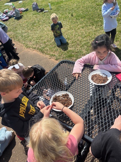 A classroom of students exploring soil outdoors A high-angle shot showing a large group of elementary students sitting in a circle on a paved outdoor area next to a grassy field. They are fully immersed in a hands-on science lesson on a bright, sunny day.  Key Details Activity: Nearly a dozen students are kneeling or sitting on the ground, each focused on white paper plates filled with soil, mulch, and organic matter. They are using their hands and small tools to sort through and examine the samples.  Environment: The scene is set outdoors at school, with green grass and backpacks visible in the background. The students are dressed in colorful casual clothes (t-shirts, sweatshirts, and patterned leggings).  Atmosphere: The image conveys a sense of active collaboration and curiosity-driven learning.