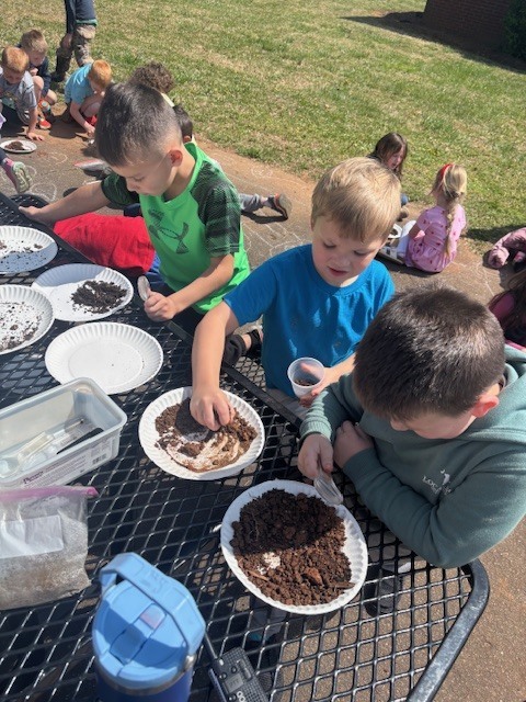 Image Description: Students examining soil samples A group of elementary-aged students are gathered outdoors at a black metal mesh picnic table on a sunny day, engaged in a hands-on science activity.  Key Details Activity: Several white paper plates containing dark soil and mulch samples are spread across the table. Three boys in the foreground are actively examining the contents; one uses a magnifying glass, another sorts through the dirt with his hands, and a third holds a small clear plastic cup.  Tools: Scientific tools are visible on the table, including magnifying glasses and a plastic bin containing droppers or pipettes.  Background: In the background, other students are sitting on the grass and a concrete walkway, also participating in the outdoor lesson. A blue water bottle is in the lower-left foreground.