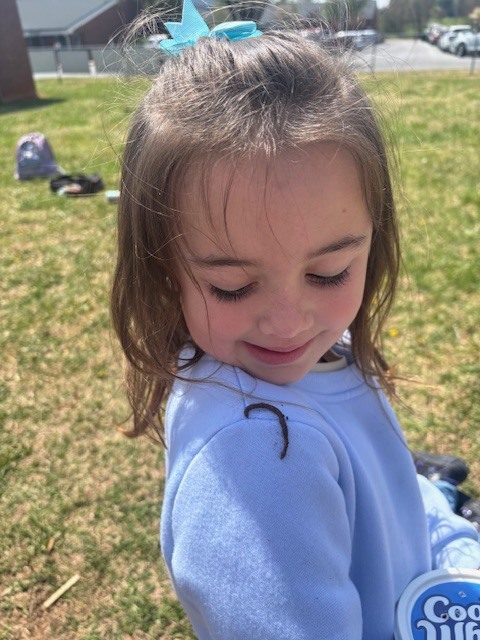 A young girl with long brown hair and a bright blue bow in her hair is smiling slightly as she looks down at her shoulder. She is wearing a soft, light blue sweatshirt.  Key Details Interaction: Resting on the girl's right shoulder is a long, thin, brown earthworm.  Environment: She is outside on a grassy area, likely a school playground or park, on a bright sunny day.  Items: In the bottom right corner, a portion of a white container with the brand "Cool Whip" can be seen.  Focus: The girl's expression and the worm on her shoulder are the main subjects of the image.