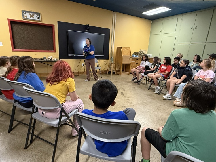 A female educator in a blue polo shirt and khaki pants stands before a group of students and adults in a classroom setting. She is holding a dark-colored animal, possibly a bird of prey or small mammal, while gesturing towards a large digital screen behind her. The students are seated in a semi-circle of gray folding chairs, focused on the presentation. The room has yellow walls, a blue ceiling, and various educational displays, including a framed butterfly collection and a specimen table. 