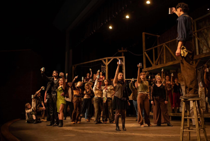 A large ensemble of student performers on a theater stage during a musical production. In the foreground, a young man stands on a wooden stepstool, holding a silver mug high in a toast. Below him and spread across the stage, approximately twenty other cast members in rustic, eclectic costumes—including vests, trousers, and a green dress—also raise silver mugs toward him. The set features a wooden balcony structure and dim, warm overhead stage lighting that creates a dramatic atmosphere.