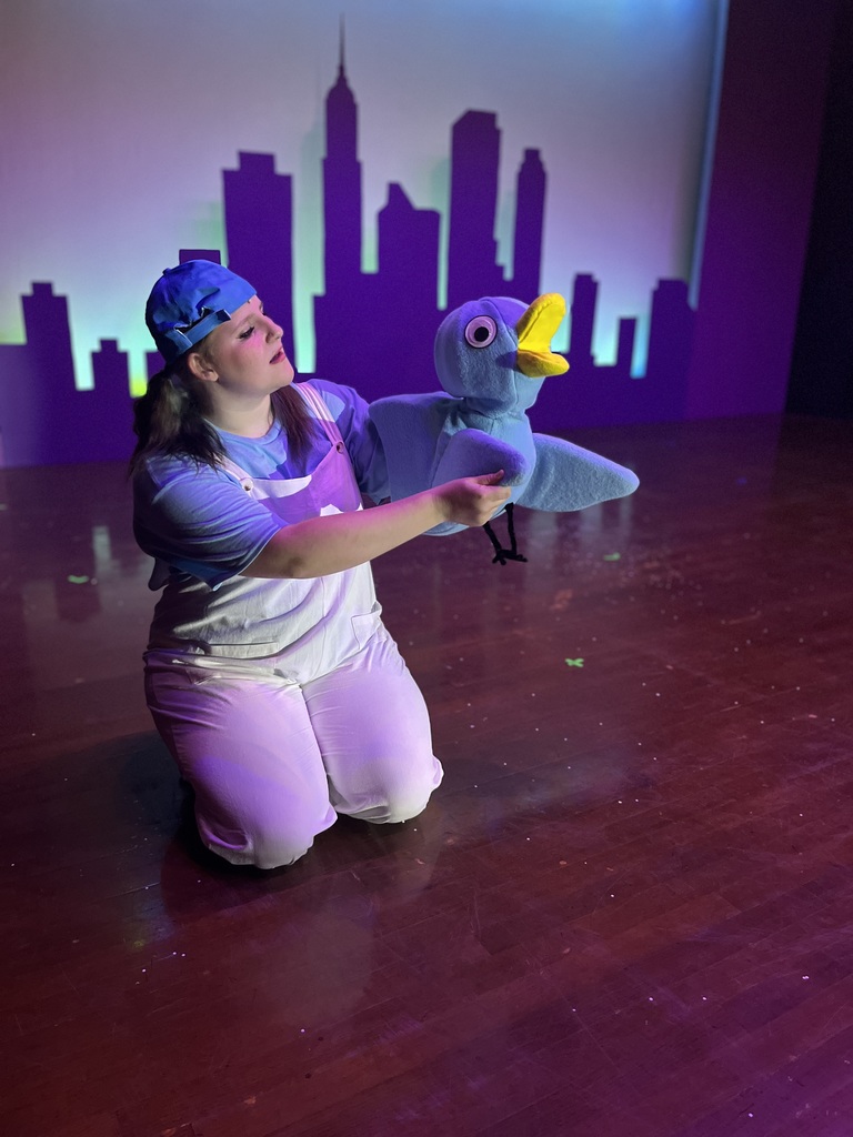 An actor kneeling on stage holding a large blue Pigeon puppet against a purple-lit city skyline backdrop.