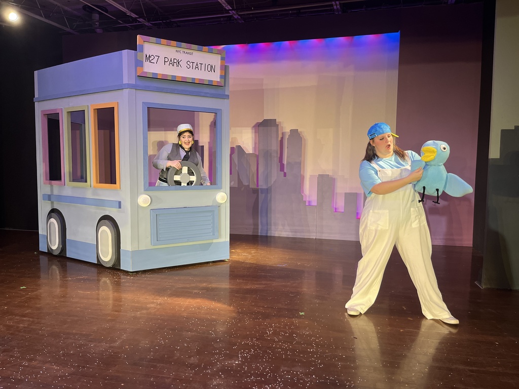 An actor at the wheel of a light blue bus scenery alongside an actor holding a large blue Pigeon puppet.