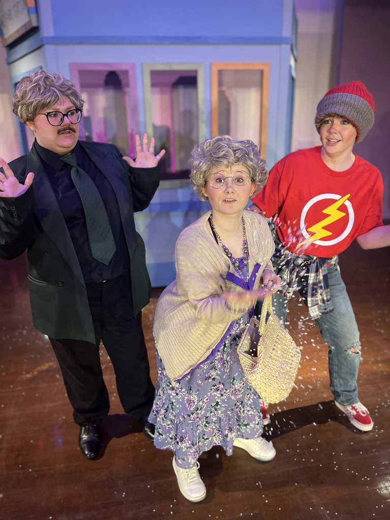 Three actors on stage: a surprised businessman in a suit, an elderly woman throwing birdseed at the camera, and a teenager in a red lightning bolt shirt.