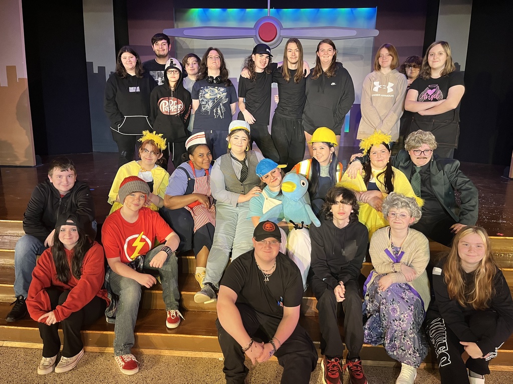 Smiling group photo of the play's cast and crew posed onstage in front of the set. The cast is dressed in their colorful costumes and the crew is dressed in black.