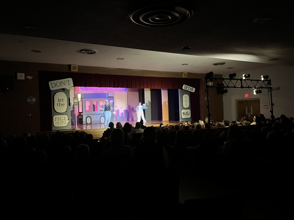 A view from the back of a darkened auditorium showing a seated audience of children watching a stage musical. The stage features the piece of bus scenery against a city skyline backdrop and a series of giant speech bubbles that read "DON'T LET THE PIGEON DRIVE THE BUS!"