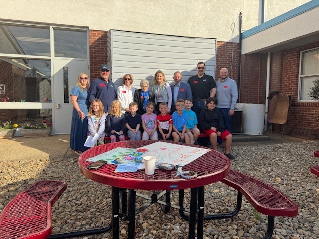 A large group of eight adults and eight children pose for a photo in a sunny outdoor courtyard. The adults stand in a row in the back, while the children are seated on a bench in front of them.  The courtyard features a ground of light-colored river stones and red metal picnic tables. On the table in the foreground, there is a large handmade "Thank You" poster decorated with colorful drawings and flowers, along with a white cup and a few personal items.  The group is positioned against the brick and siding of a school building. Many of the adults are wearing the same red nametags seen in previous photos, suggesting a community dedication or "thank you" event for the Union Courtyard, likely involving the donors and students.
