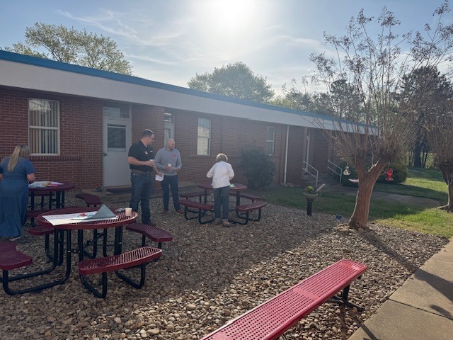 Four people are gathered in an outdoor courtyard area of a brick school building on a sunny day. The courtyard features a ground cover of light-colored river stones and several circular red metal picnic tables with attached benches.  To the left, a woman in a long denim dress stands by a table holding papers.  In the center, two men in casual attire (one in a black polo and one in a grey quarter-zip) stand near another table, talking and holding coffee cups.  A woman in a white shirt and dark pants stands with her back to the camera at a central table, appearing to organize materials.  In the foreground, a long red bench sits alongside a concrete walkway. A small, budding tree stands to the right, and the sun shines brightly from the upper center of the frame, casting long shadows across the gravel.