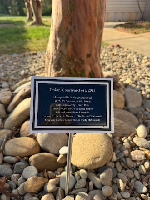 A close-up of a commemorative plaque mounted on a metal stake, situated in a bed of smooth river stones at the base of a tree. The plaque is black with a silver border and white text.  The text on the plaque reads:  Union Courtyard est. 2025  Made possible by the generosity of:  RE/MAX Crossroads—Will Poteat  Wise Landscaping—David Wise  Turner Houser Insurance—Jamie Houser  Edward Jones—Brent Reynolds  Barbara S. Houser in Memory of Katherine DiBernardo  Chip and Camille Sain in Honor Shelly McConnell  In the background, the sun casts a warm glow on the tree trunk and the surrounding courtyard area.