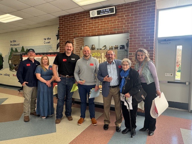 A group of seven people stands together for a photo in a school hallway. From left to right: a man in a black baseball cap and navy button-down, a woman in a long denim dress, a tall man in a black polo shirt and jeans, a man in a grey quarter-zip sweater, a man in a blue patterned blazer, an elderly woman in a black coat holding a cane, and a woman in a lavender top and grey vest.  Most of the individuals are wearing red nametags. They are positioned in front of a brick wall featuring a built-in trophy case and a digital clock displaying 9:16 AM, Mon, Mar 30. To the left, a mural on the wall includes the text "to the Piedmont" and "to the Coast." The group appears to be at a community or school event, with some holding papers or folders.