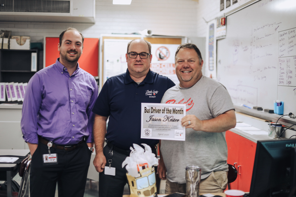 Three men stand in a classroom, smiling, with one holding a certificate reading "Bus Driver of the Month."