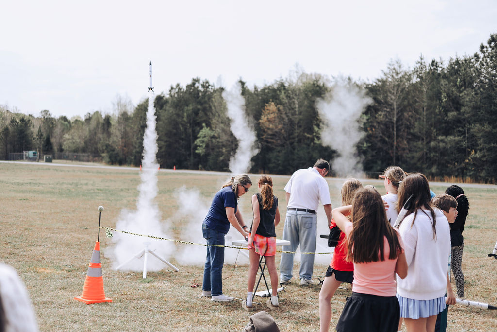 Several people watch two rockets being launched in an open field with a forest background.