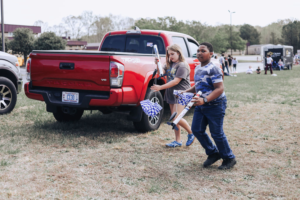 Two people near a red pickup truck hold foam bats and appear to be walking toward the camera.