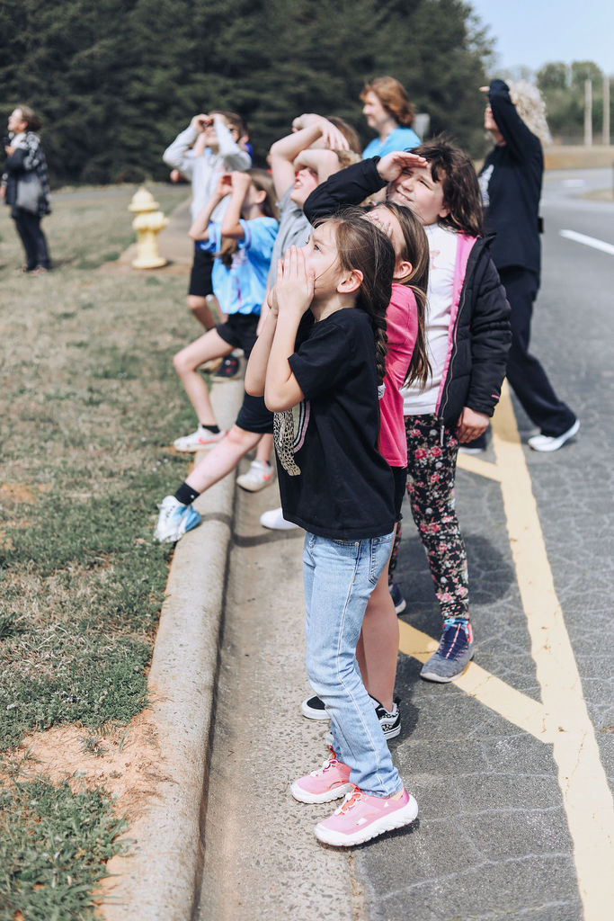 Children line up on a curb, watching intently through binoculars at something in the distance.