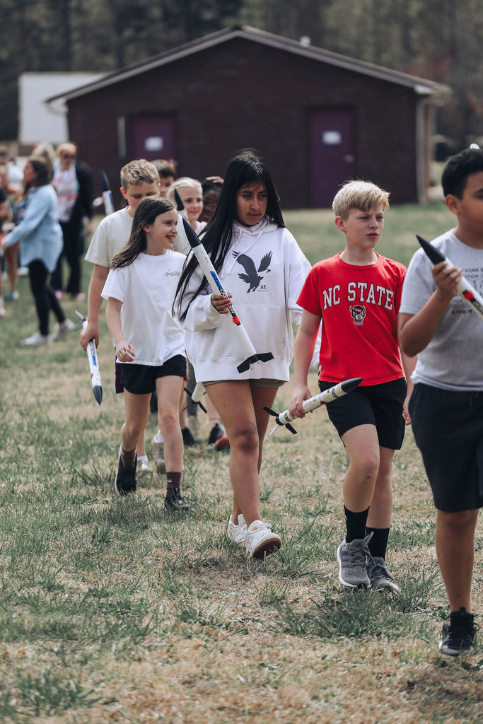 Group of children, possibly on a field trip, walking in a line on grass, carrying toy rocket launchers.