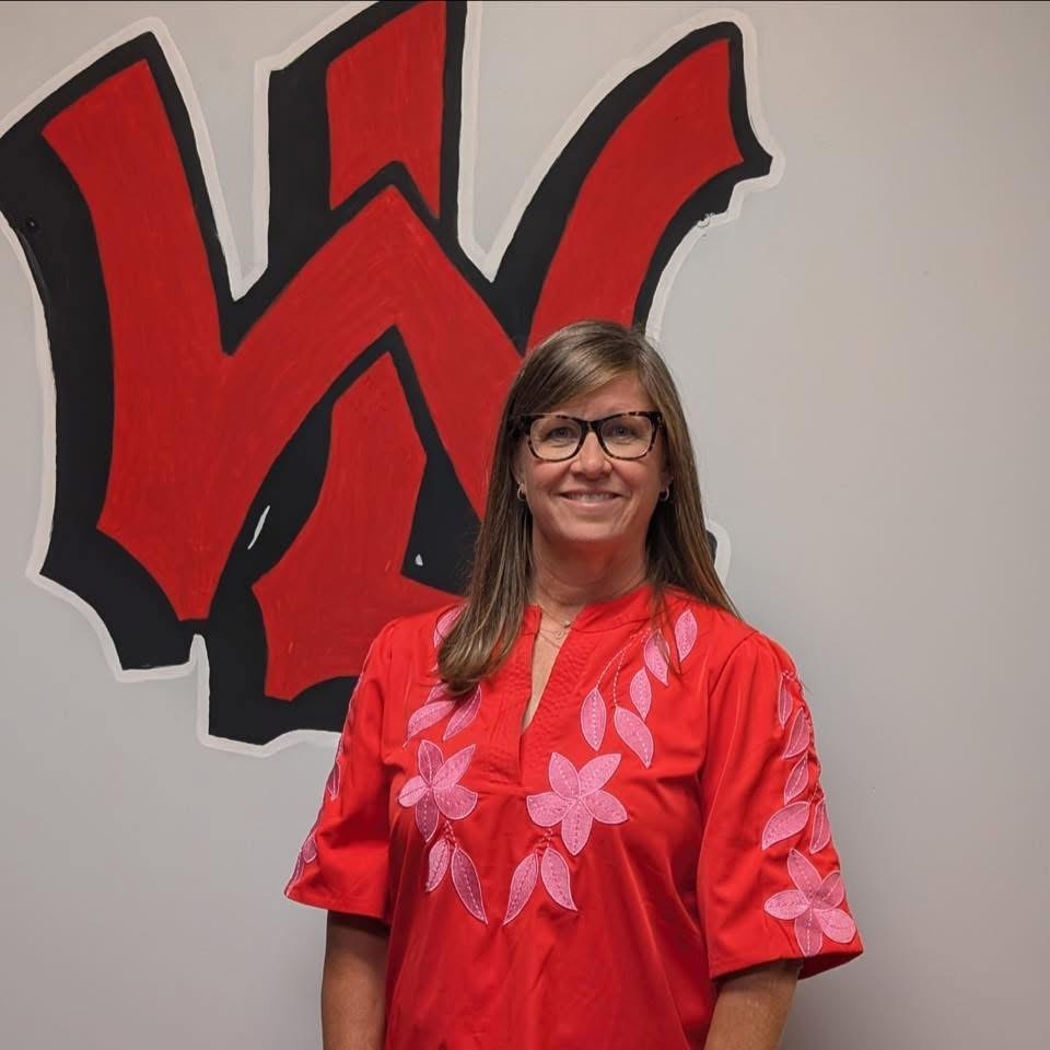 A woman in a red blouse with flower patterns stands in front of a white wall with a red W.