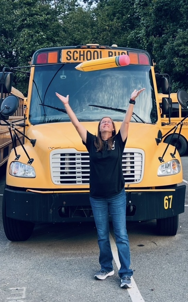 A woman in a black shirt and jeans stands with arms raised in front of a yellow school bus.