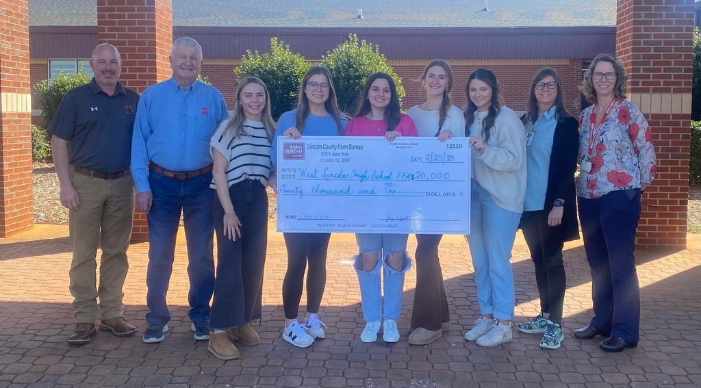 People in casual clothing standing next to a brick wall holding a large check.