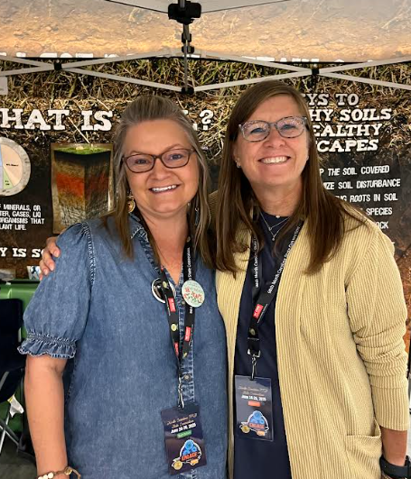 Two women smile at the camera, standing next to an educational display with text about soil health.