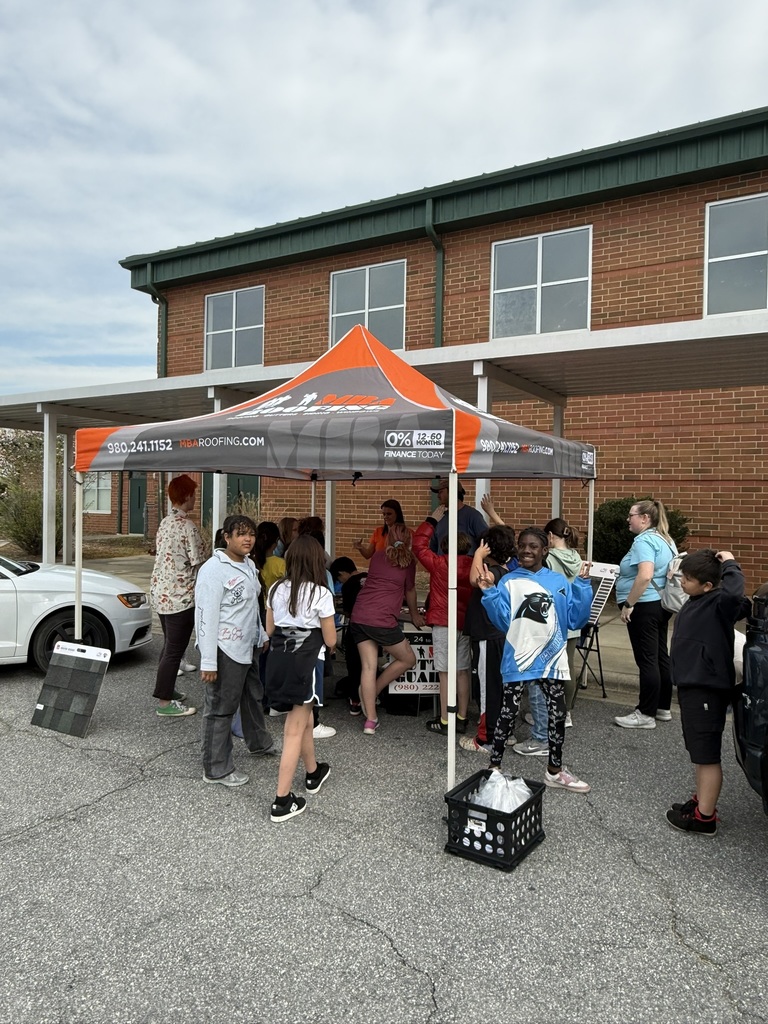 A group of people gathered under a tent with a white car parked nearby in front of a brick building.