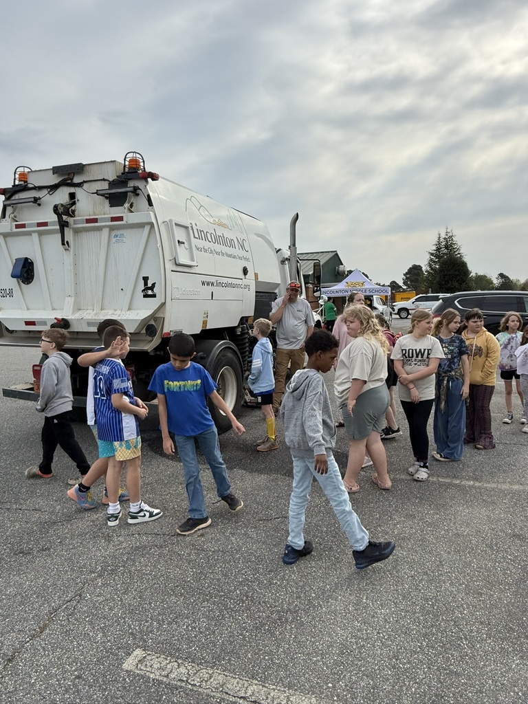 A group of people, including children, are gathered in a parking lot, with a white trash truck nearby.