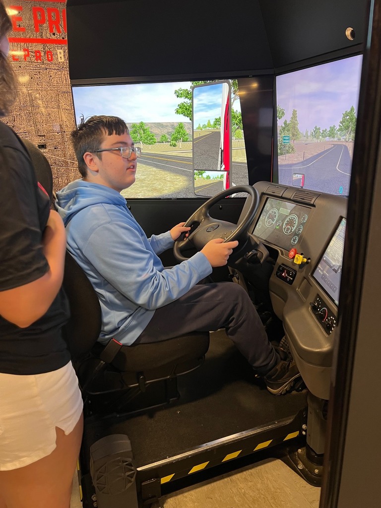 A person with glasses drives a vehicle simulator while another person in shorts stands nearby.