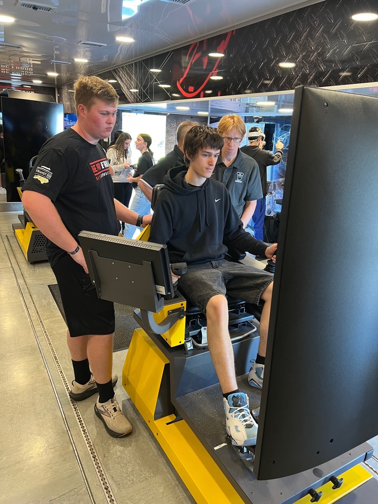 A person in a hoodie and sneakers uses a large screen monitor while another person stands behind.