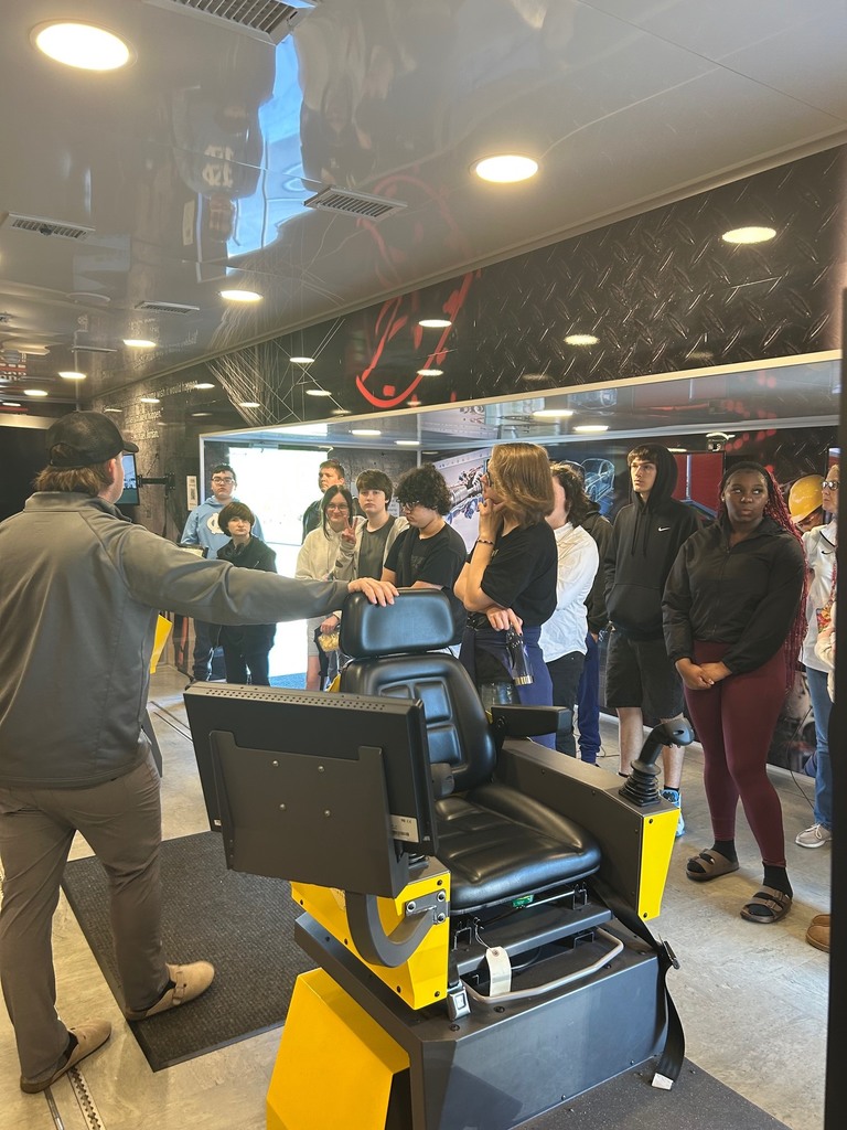 People stand around a chair simulator, with one person gesturing. The room has black walls and white ceiling with lights.
