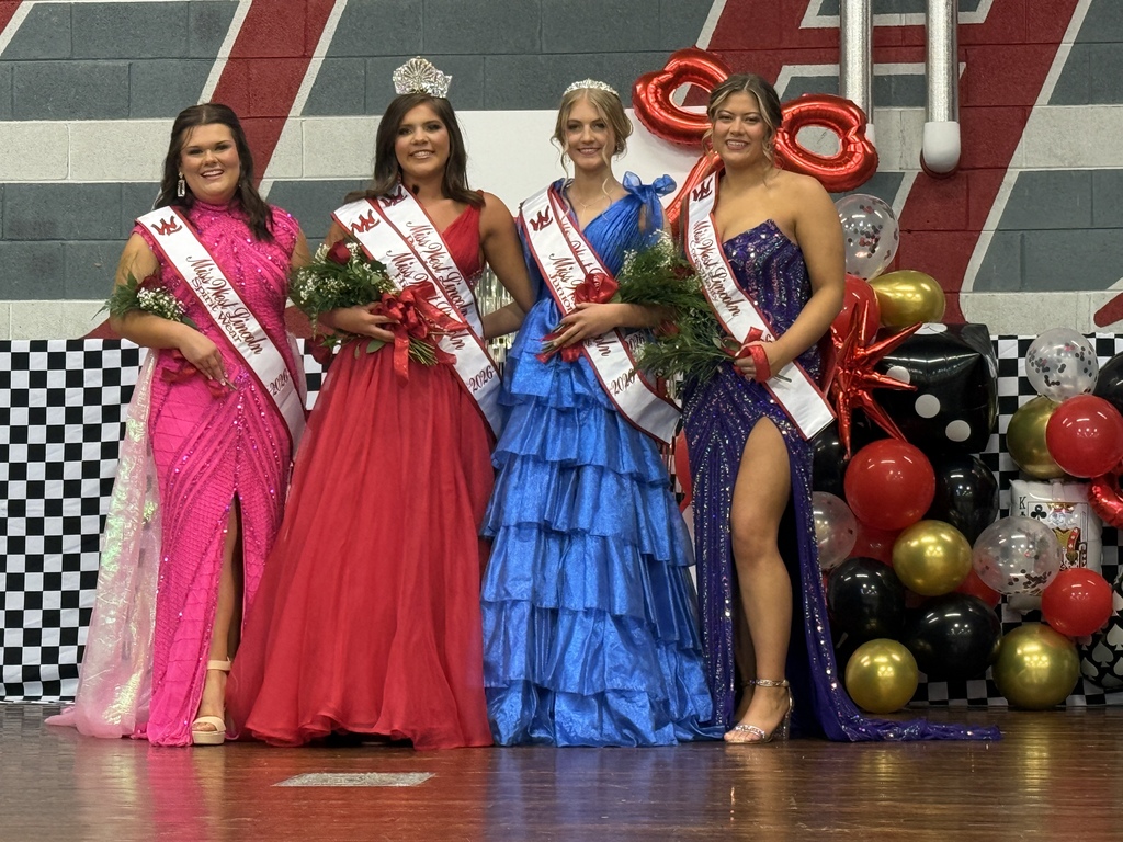 Five women in gowns, sashes, and crowns stand on a stage. Each holds flowers. Behind them, red and white striped walls, balloons, and a checkered floor.