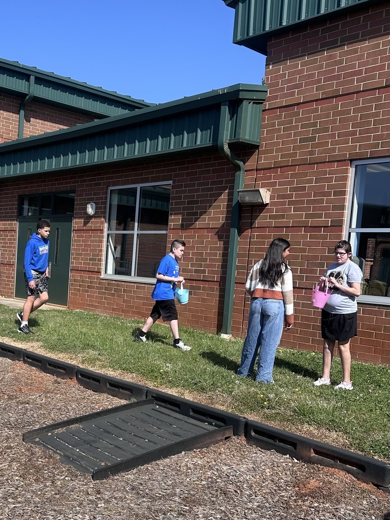 Four people outside a brick building, one holding a bucket, others carrying baskets. One person running, others standing on grass.