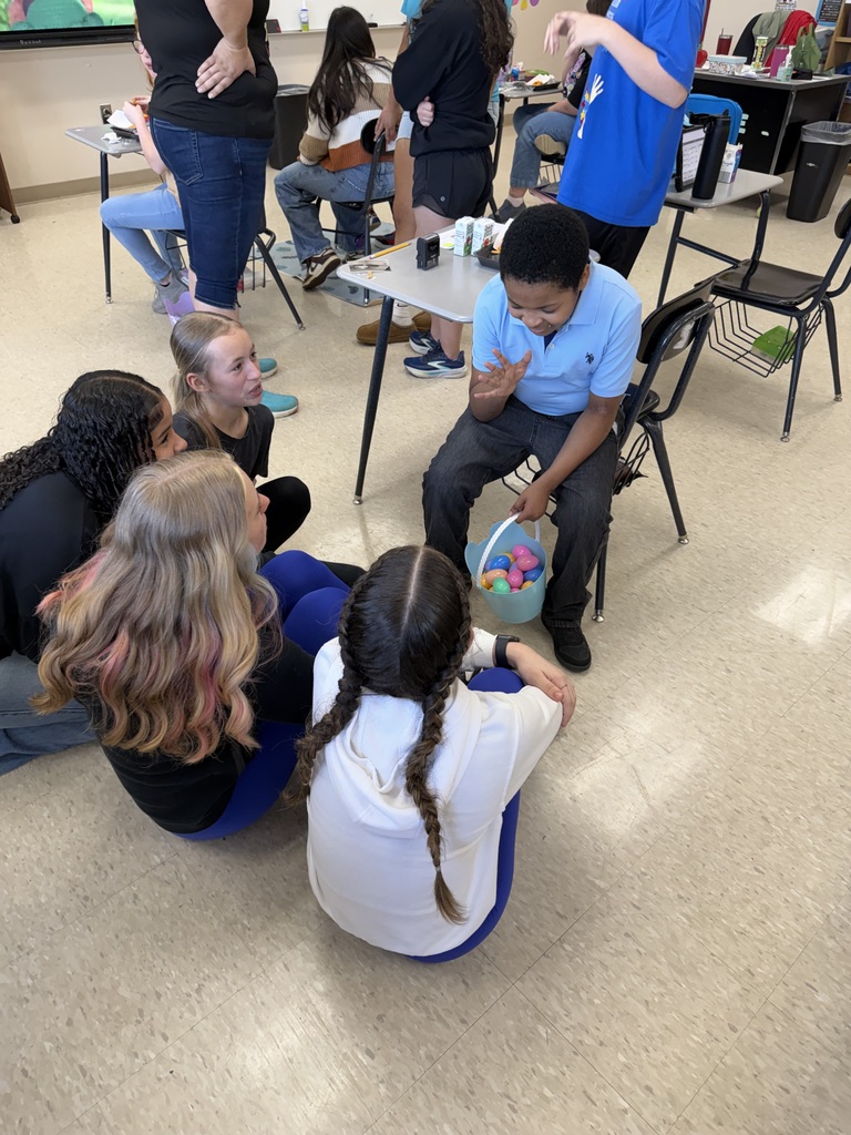 Several students are seated on the floor in a classroom. A person in a blue shirt holds an Easter basket.