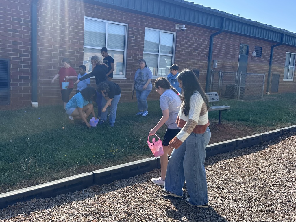 Multiple individuals gather around a brick building, some crouched on the grass, holding pink buckets. One individual stands, holding a pink bucket.