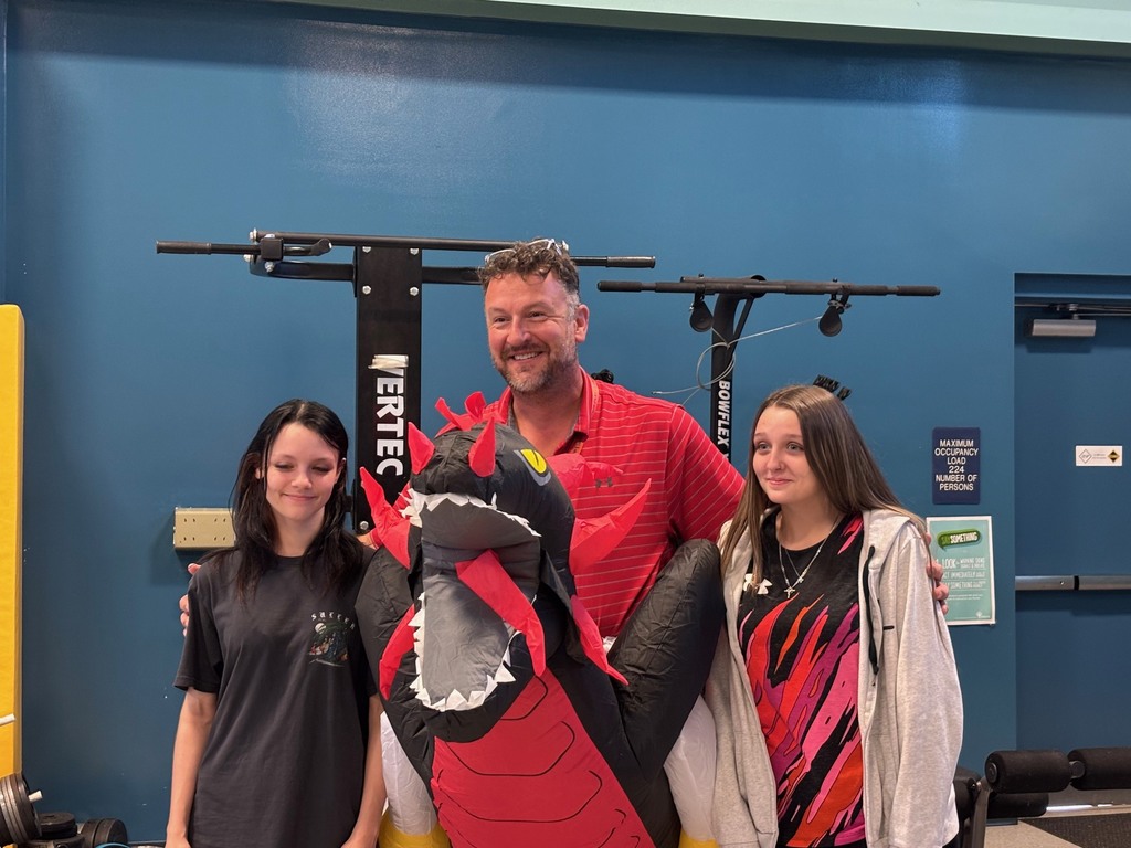 Three people, including a man and two women, stand together in a gym. The man is in a red shirt.